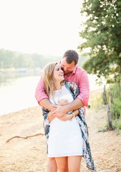 a man and woman hugging each other on the beach near water with trees in the background