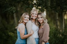 three beautiful women standing next to each other in front of some trees and bushes with their arms around one another