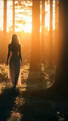 a woman in a white dress walking through the woods at sunset with her back to the camera