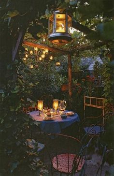 an outdoor dining area is lit up with candles and lanterns in the evening time, surrounded by greenery