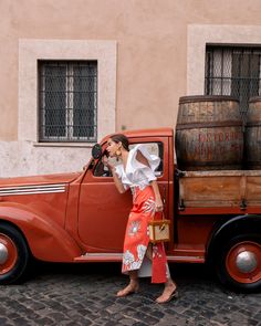 a woman standing next to an old red truck with barrels on the back and a barrel in the trunk