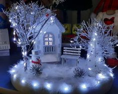 a small white house with lights and trees in the snow next to a teddy bear