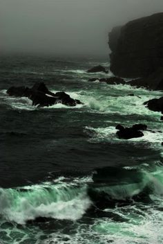 the ocean is green and choppy as it comes towards shore with large rocks in the foreground