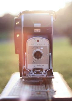 an old camera sitting on top of a wooden table in the sun light, with grass and trees behind it