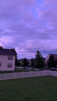 a white picket fence in front of a house at dusk with purple clouds above it