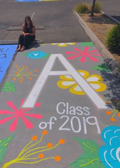 a girl sits on the sidewalk painted with flowers and letters that spell out class of 2019