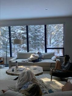 two people sitting on couches in front of large windows with snowy trees outside the window