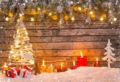 a christmas tree with lights and presents in the snow next to a wooden fence covered in snow