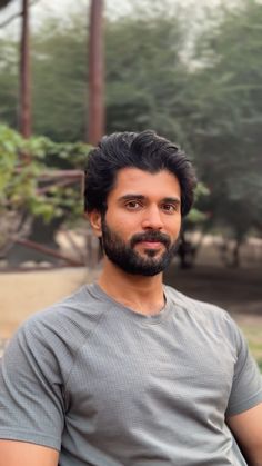 a man with a beard and grey shirt posing for a photo in front of some trees