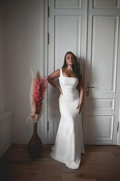 a woman in a white dress standing next to a vase with pink feathers on it