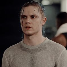 a man with long hair wearing a grey sweater and looking at the camera while standing in front of other people