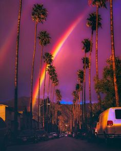 a rainbow in the sky over palm trees and cars parked on the side of the road