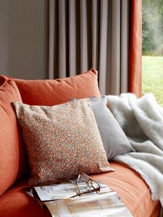 an orange couch with two pillows and a magazine on it's side, in front of a window