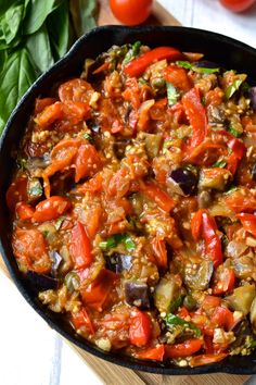 a skillet filled with vegetables on top of a wooden cutting board next to tomatoes and basil