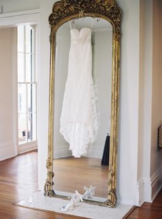 a wedding dress is hanging in front of a large gold mirror with shoes on the floor