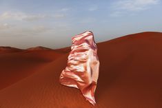 a pink blanket is in the middle of an orange sand dune, with blue sky and clouds above it