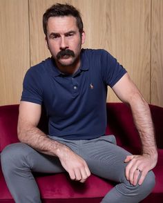 a man sitting on top of a red couch wearing grey pants and a blue polo shirt