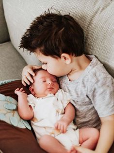 a young boy is holding his sleeping baby on the couch while he looks down at him