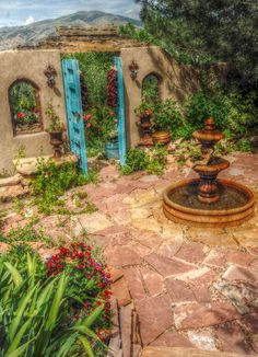 a painting of an outdoor fountain surrounded by greenery and potted plants with blue doors in the background