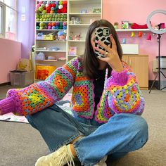 a woman sitting on the floor with her cell phone up to her face while wearing a colorful crocheted jacket