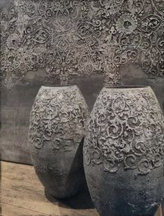 two gray vases sitting on top of a wooden table next to a wall with an intricate design