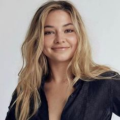 a woman with long blonde hair wearing a black shirt and smiling at the camera while standing in front of a white background