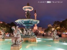 the fountain is lit up at night with many statues around it and people walking around