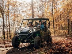 four people riding in the back of a green vehicle on a dirt road surrounded by trees