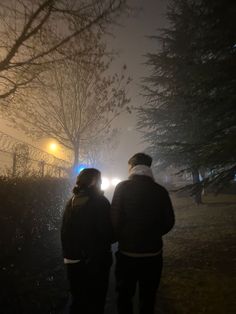 two people walking down a sidewalk at night in the fog with street lights behind them