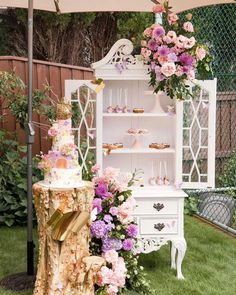 a table topped with a cake next to a white cabinet filled with pink and purple flowers