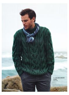 a man wearing a green sweater and scarf standing next to the ocean with his hands in his pockets