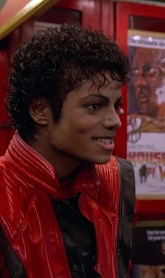 a young man with curly hair wearing a red leather jacket and black shirt in front of a movie poster