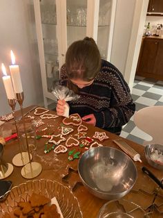 a woman decorating gingerbreads on a table with candles and other baking supplies