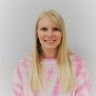 a woman with long blonde hair wearing a pink and white shirt smiling at the camera