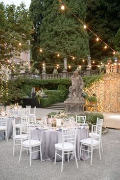 an outdoor dinner table with white chairs and lights strung from the trees in front of it