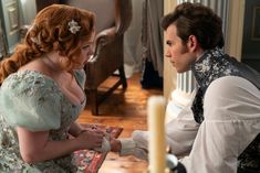 a man and woman dressed in period clothing sitting at a table looking at each other