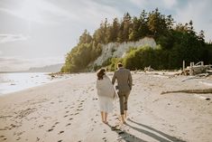 a man and woman walking on the beach holding hands with trees in the background at sunset