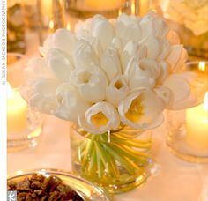 white flowers and pecans in a glass vase on a table with candles behind it