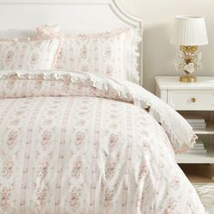 a white bed with pink flowers and ruffles on the comforter is next to a night stand