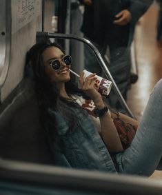 a woman wearing sunglasses and drinking from a carton while sitting on a subway train