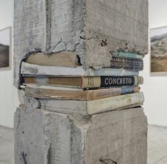 a stack of books sitting on top of a cement pillar in an art gallery next to paintings