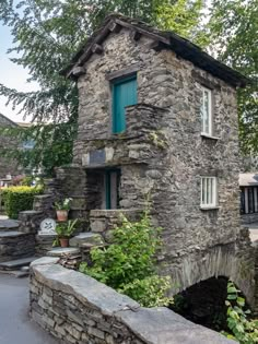 an old stone building with green shutters next to a small bridge and trees in the background