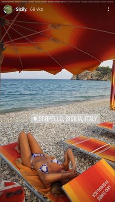 a woman laying on top of a beach under an orange and red umbrella next to the ocean