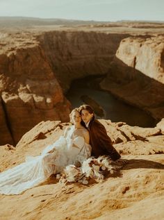 two people sitting on the ground in front of some rocks and water, one person is holding her head