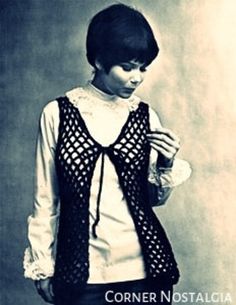 a woman standing in front of a wall wearing a black vest and white shirt with crocheted details