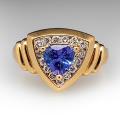 This colorful contemporary ring is centered with one (1) triangular cut natural tanzanite set into a three-prong setting. The tanzanite is bordered with fifteen (15) channel set round brilliant cut diamonds. The ring measures 13.2mm at the top, rises 7.2mm above the finger, tapering to 2.3mm wide and 0.7mm thick at the base of the shank. The ring is currently a size 7. Tanzanite Diamond Ring, Tanzanite Diamond, Contemporary Ring, Natural Tanzanite, Channel Set, One 1, Round Brilliant Cut Diamond, Brilliant Cut Diamond, Round Brilliant