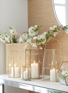 three gold lanterns with white flowers and candles on a mantle in front of a mirror