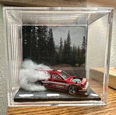 a red toy car in a clear case on a wooden table with pine trees behind it