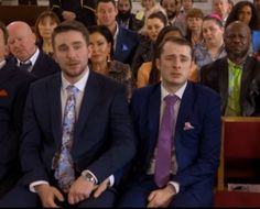 two men sitting next to each other in front of a group of people wearing suits and ties