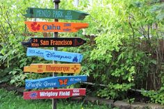 a sign post with many different colored signs on it in front of some bushes and trees